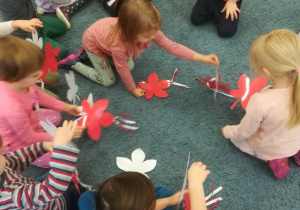 Dzieci na dywanie układają girlandę – biały kwiatek na górze, czerwony na dole.